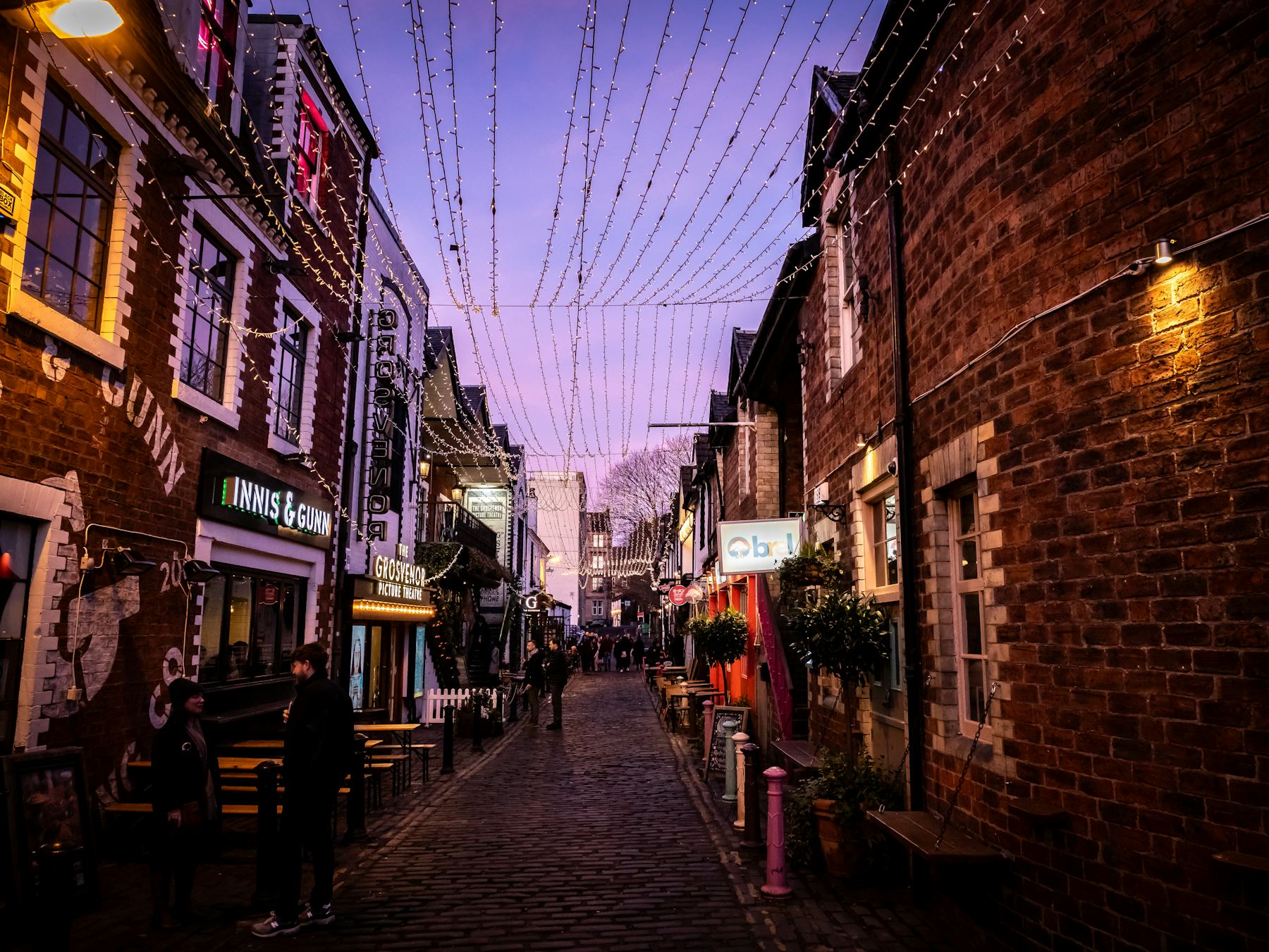 A brick-lined street hung with lights, storefronts glowing at dusk.