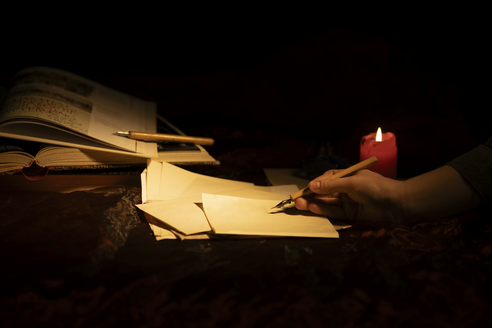 A hand writing on paper by candlelight at a dark desk.
